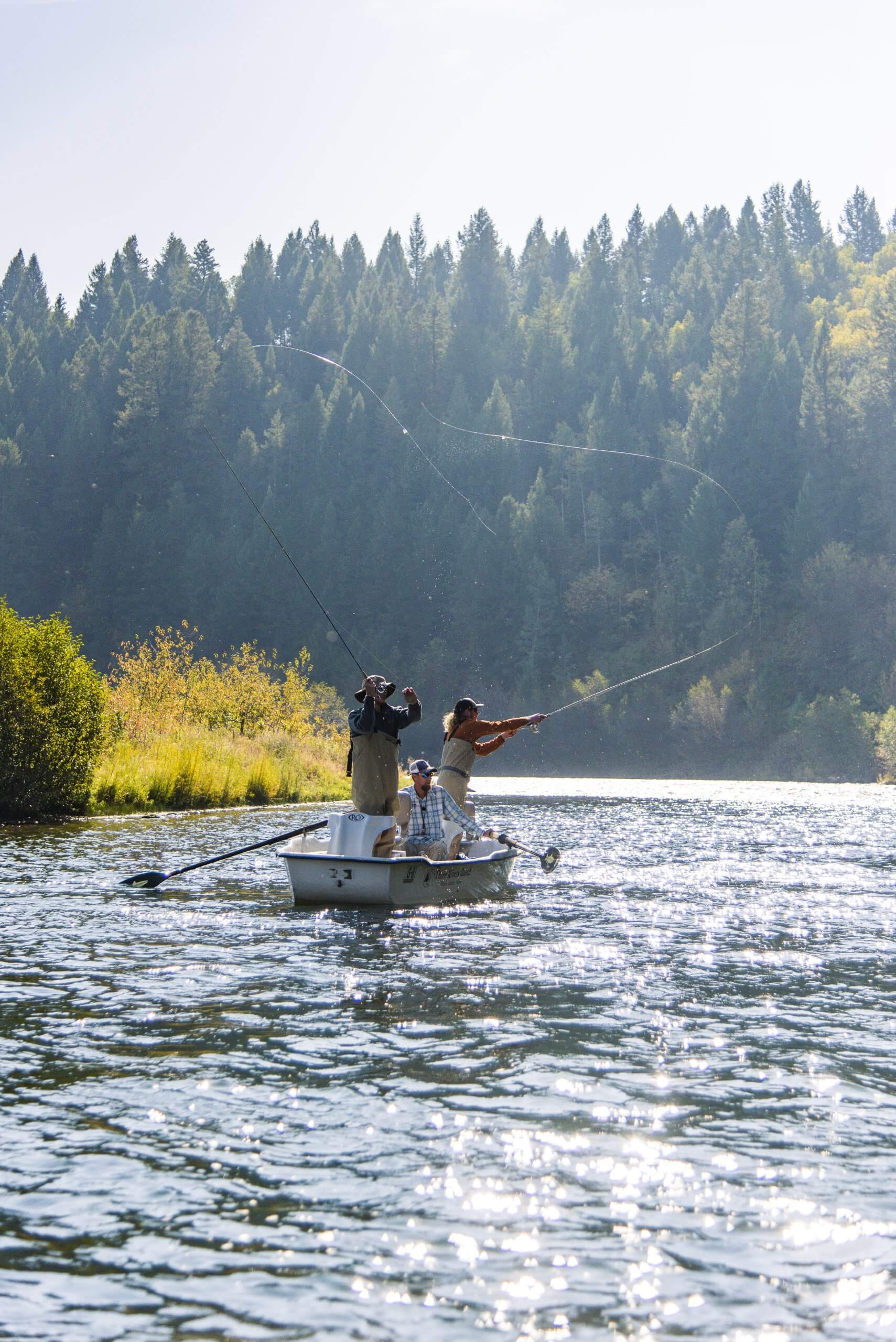 Three people fly fishing in a small boat on Henry's Fork of the Snake River, with a forest in the background.
