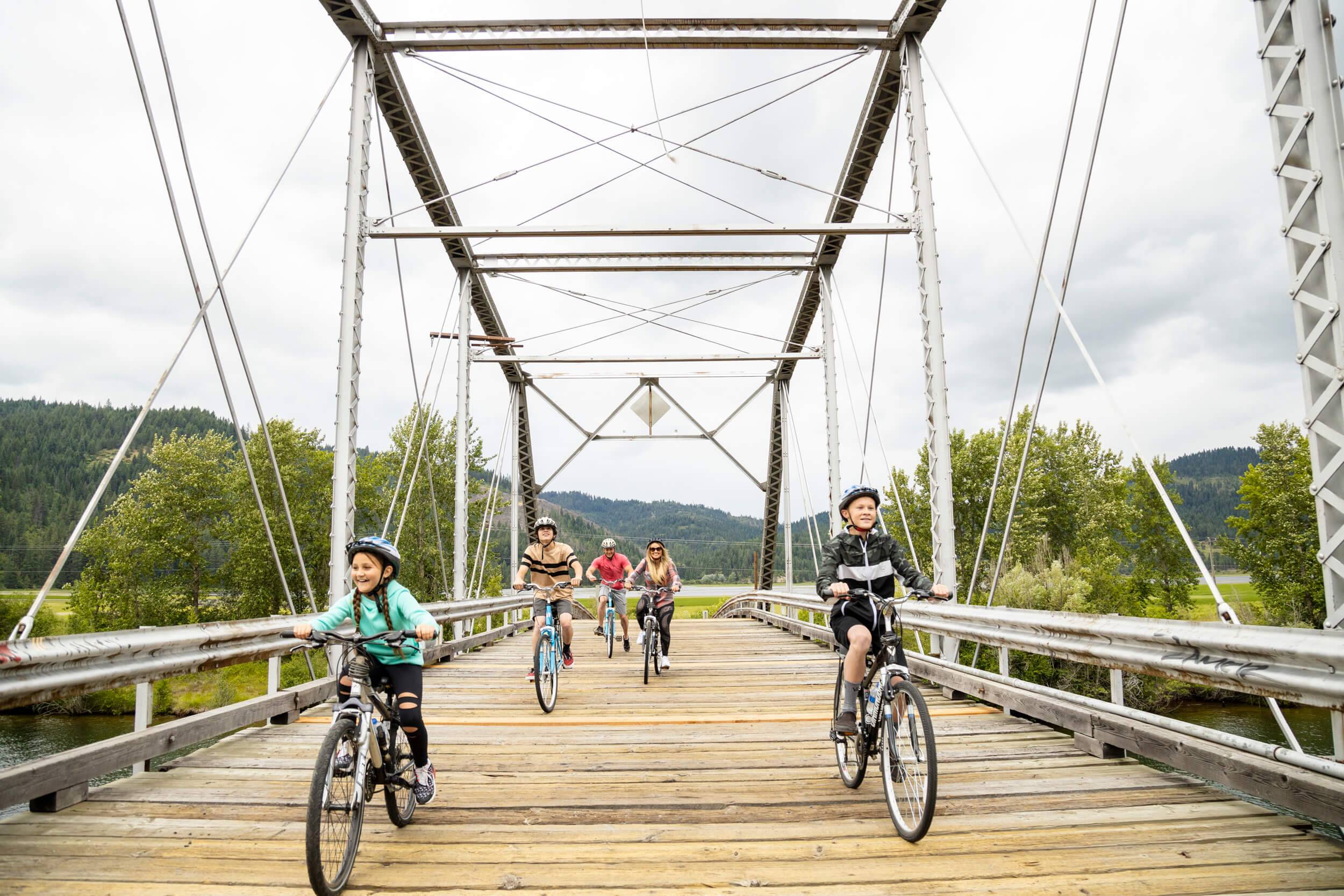 A family of five rides bikes over a wooden bridge along the Trail of the Coeur d’Alenes.
