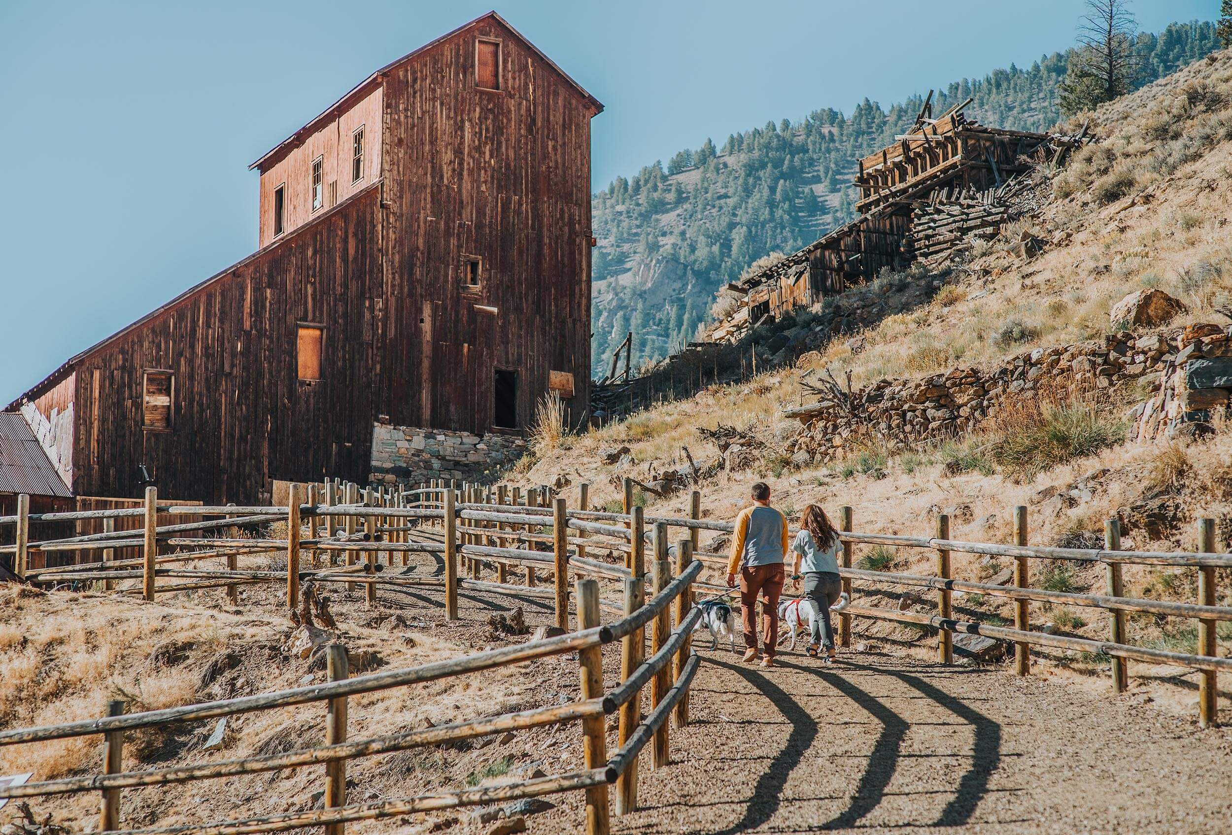 A couple walking with their two dogs along a dirt road towards a tall historic wooden building.