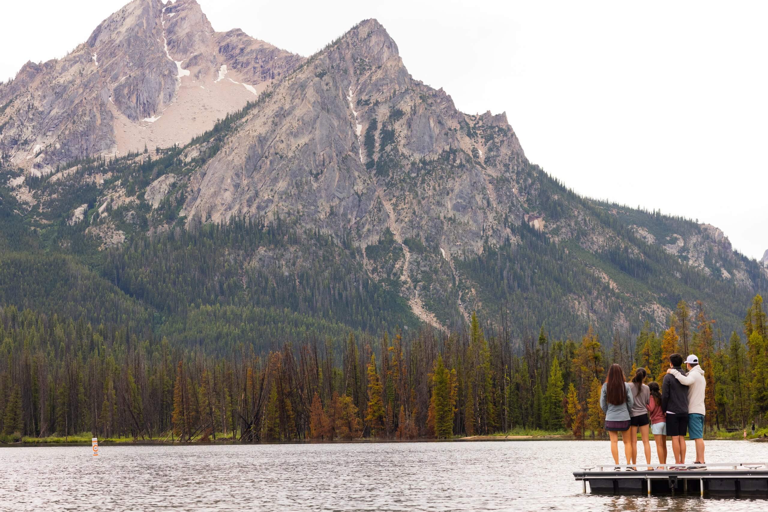 a group of people standing on a dock overlooking Stanley Lake with a forest of trees and mountains in the background
