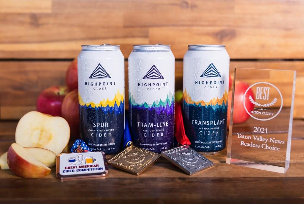 Three cans of Highpoint Cider positioned next to awards and an apple.