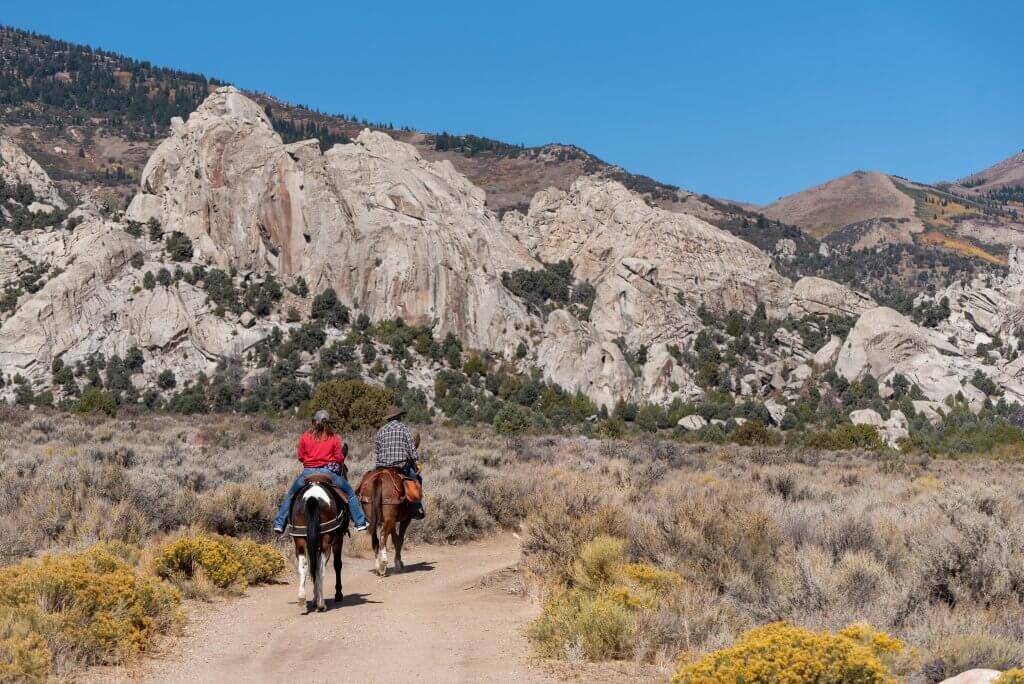 two horseback riders on dirt trail riding away with rugged rock formations in the distance at Castle Rocks State Park.