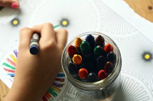 top view of a glass jar of crayons sitting on top of a coloring sheet and a child's hand holding a crayon