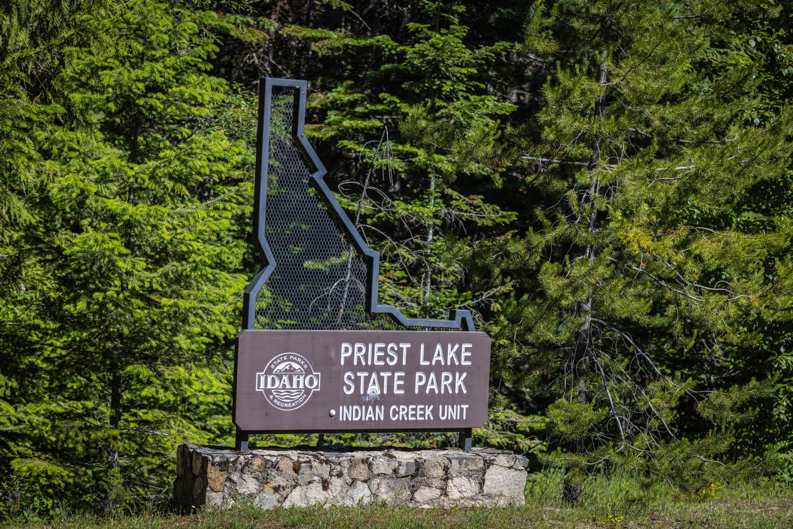 A metal sign of the outline of Idaho that reads "Priest Lake State Park Indian Creek Unit," with a forest in the background.