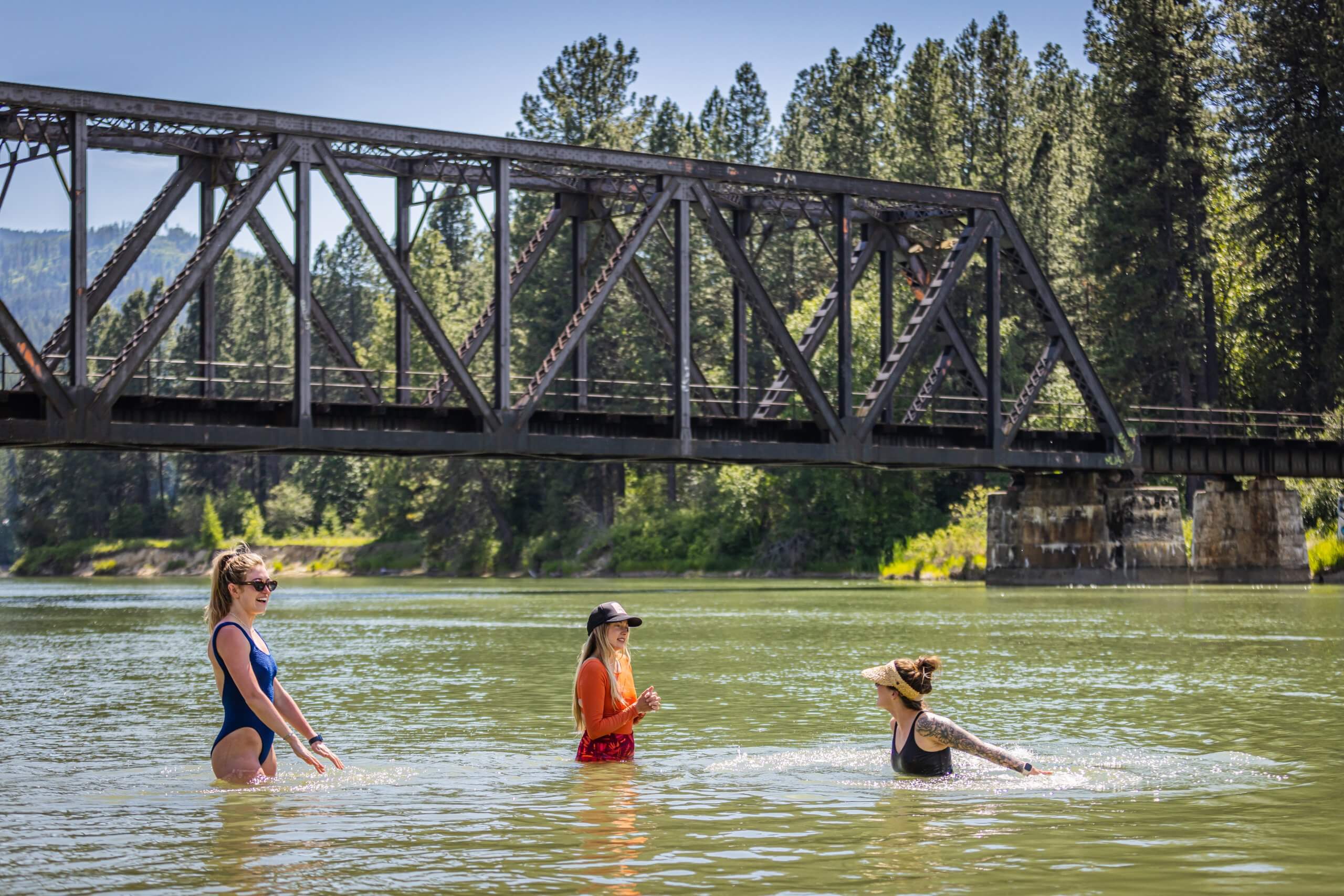 Three women wading in Priest River with a bridge and trees in the background.
