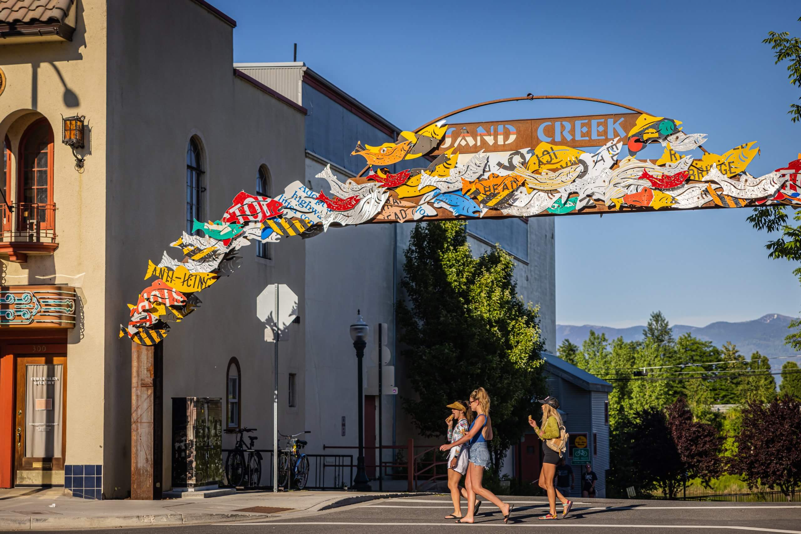 Three women walk across a street under a colorful sign that reads Sand Creek.