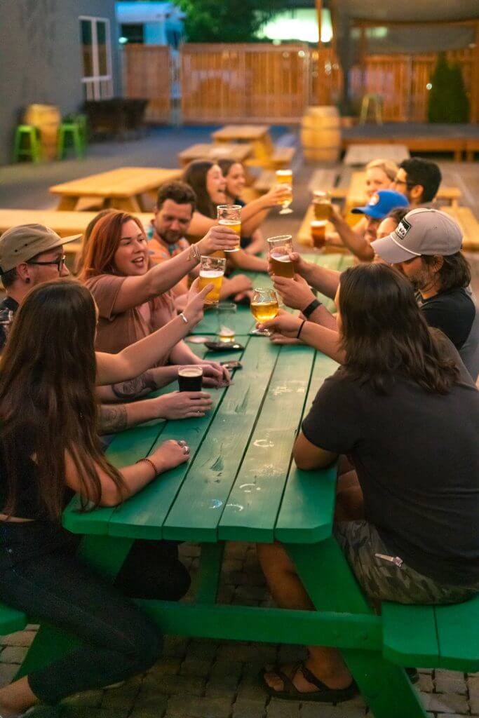 A group of people cheers glasses of beer from Lost Grove Brewing at a picnic table in their outdoor patio area.