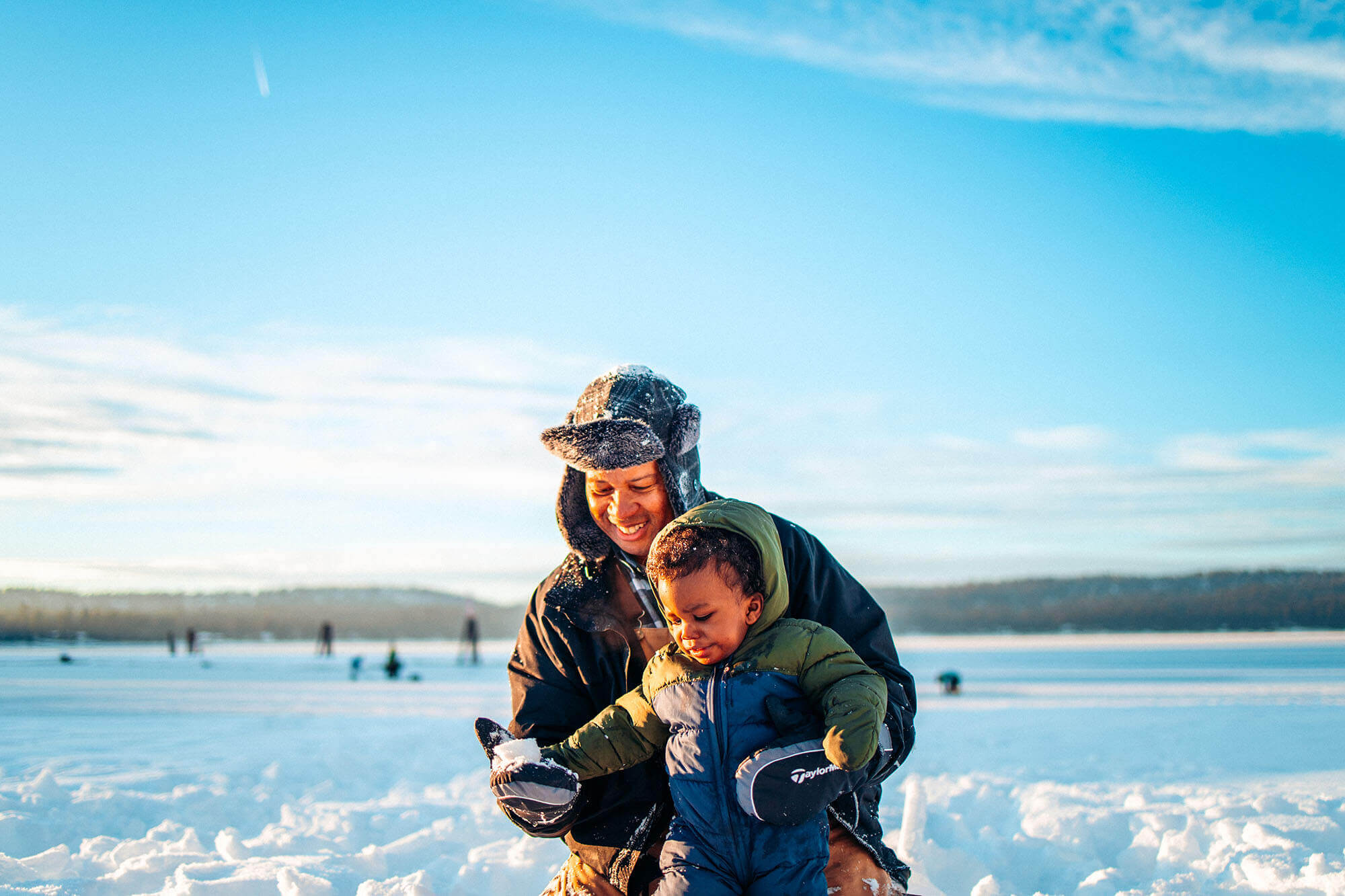 A person and child in cold weather gear play in the snow on Payette Lake under a blue sky.
