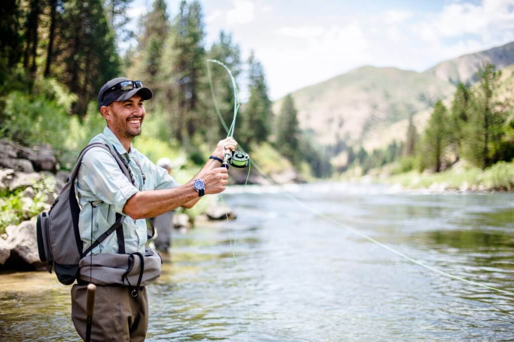 A smiling man in a hat casting a fishing line into the river.