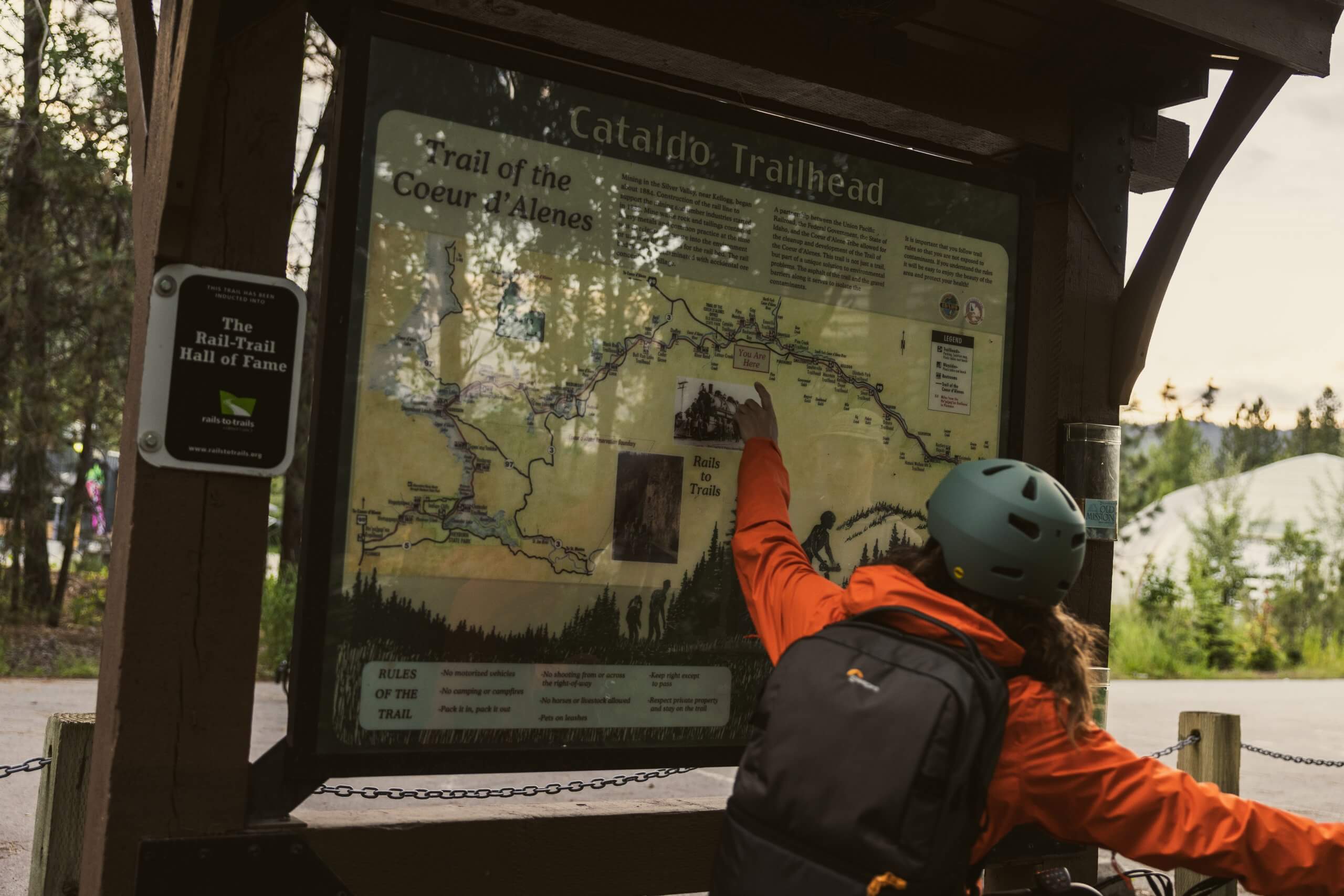 A rider points at a sign displaying a map of the Cataldo Trailhead, located at the trailhead.