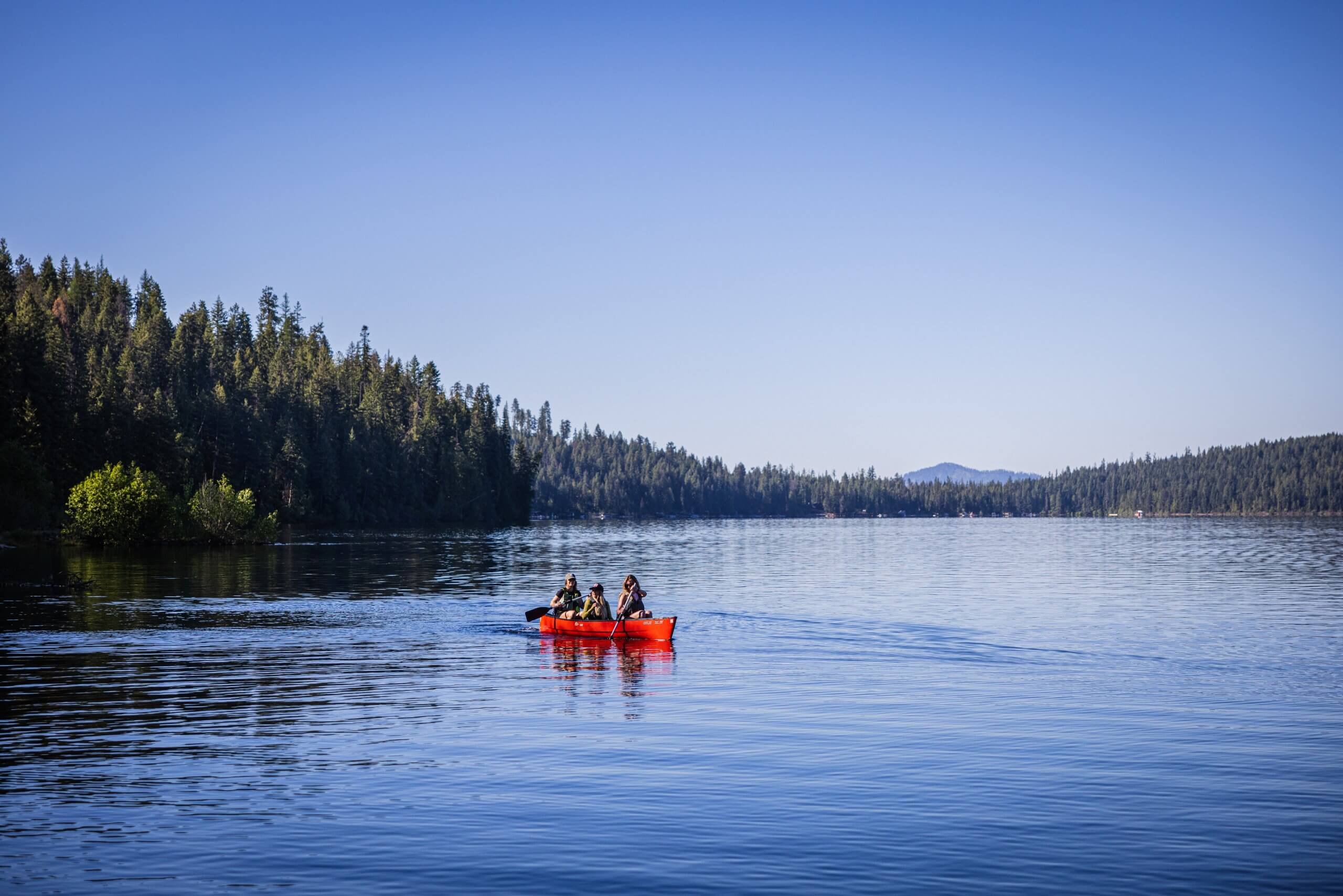 Three women paddling a red canoe across tree-lined Priest Lake at Priest Lake State Park.