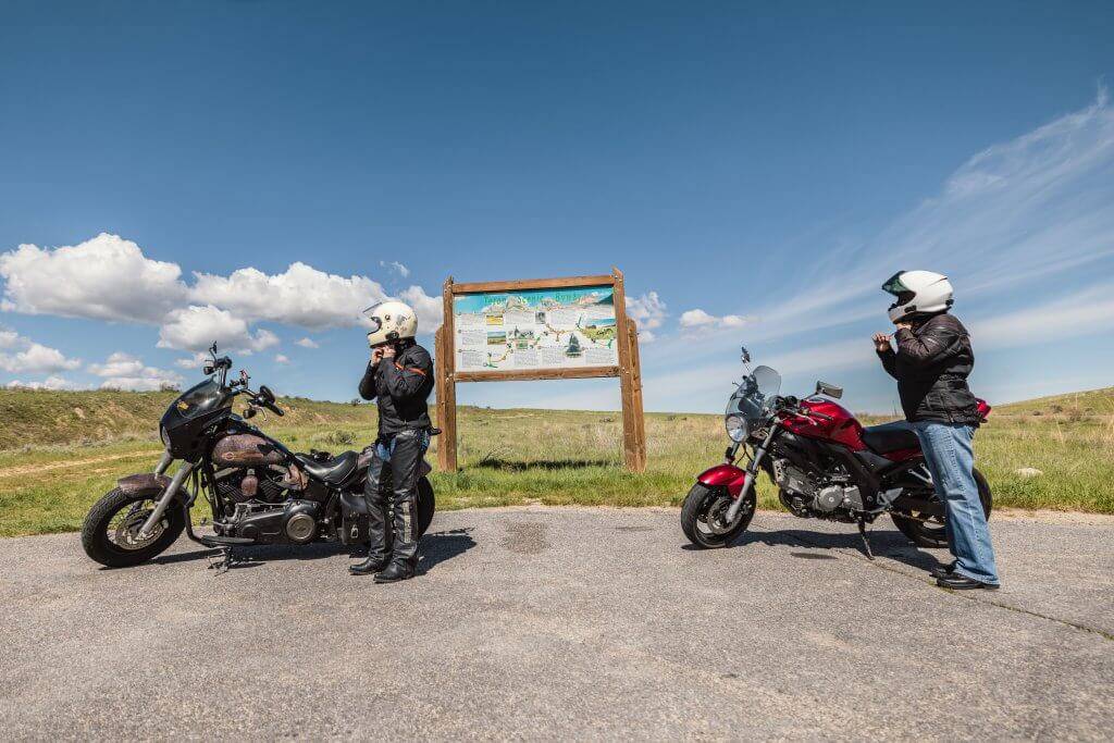 two motorcycle riders in helmets standing next to Teton Scenic Byway signs with blue skies in the background