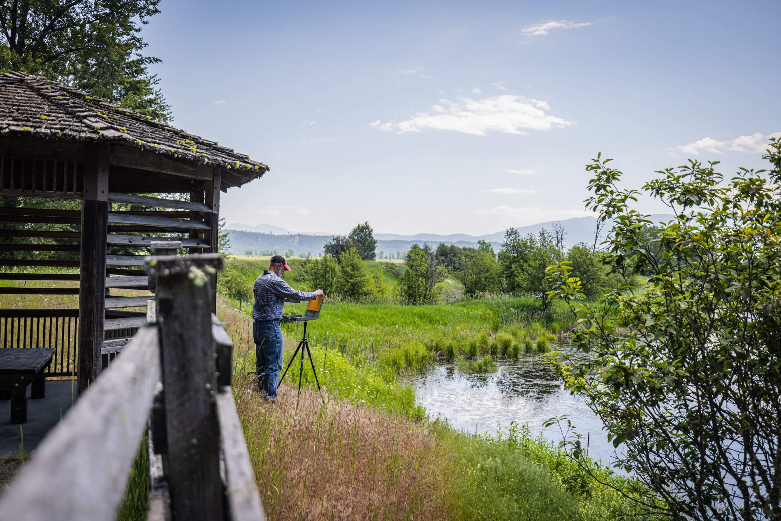 A man plein air painting in a field of tall grass in front of a body of water at Kootenai National Wildlife Refuge.