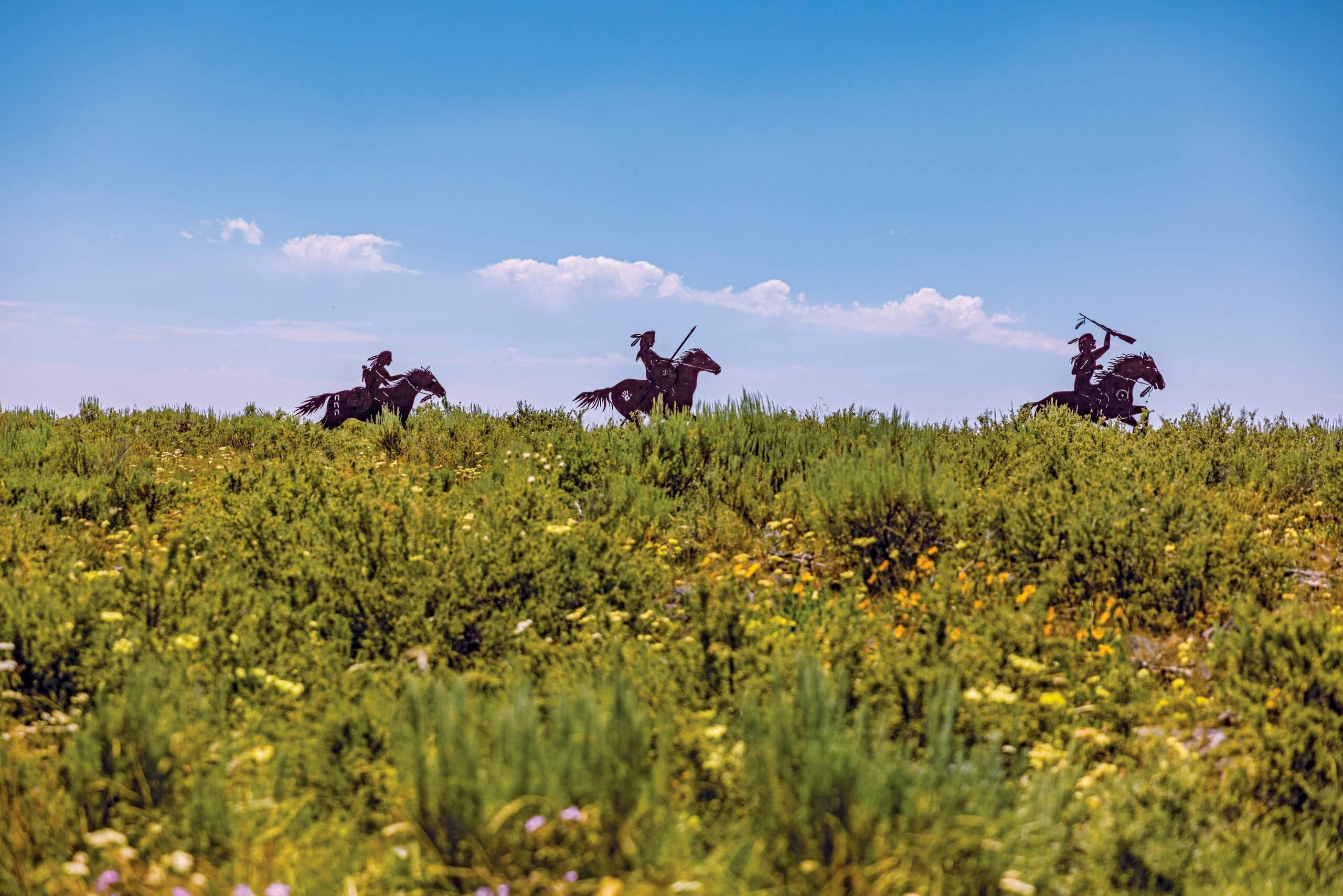 metal silhouettes of three nez perce warriors riding horses at the top of a hill covered in tall grass and wildflowers at camas meadows battle site.