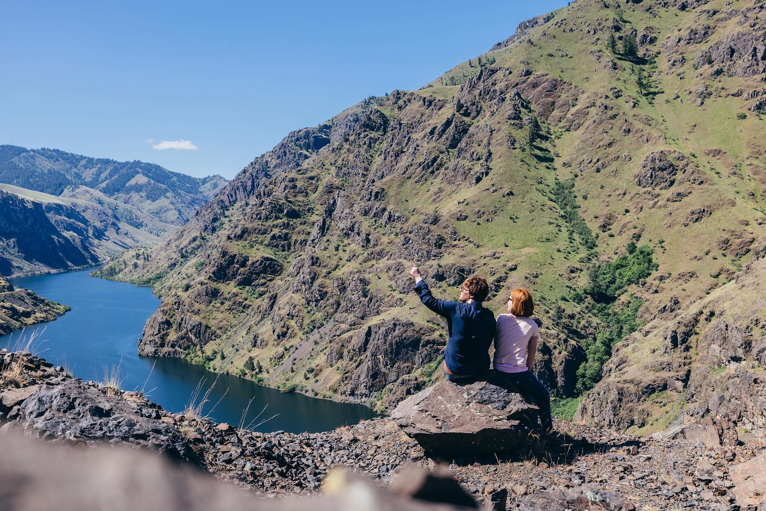two people sitting on a rock looking out over a river running through a canyon