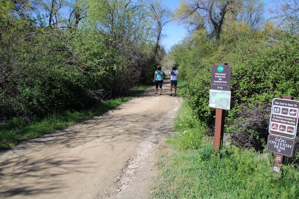 Two people walk on the packed gravel Hulls Gulch Trail in the Boise foothills surrounded by trees and shrubbery on each side.