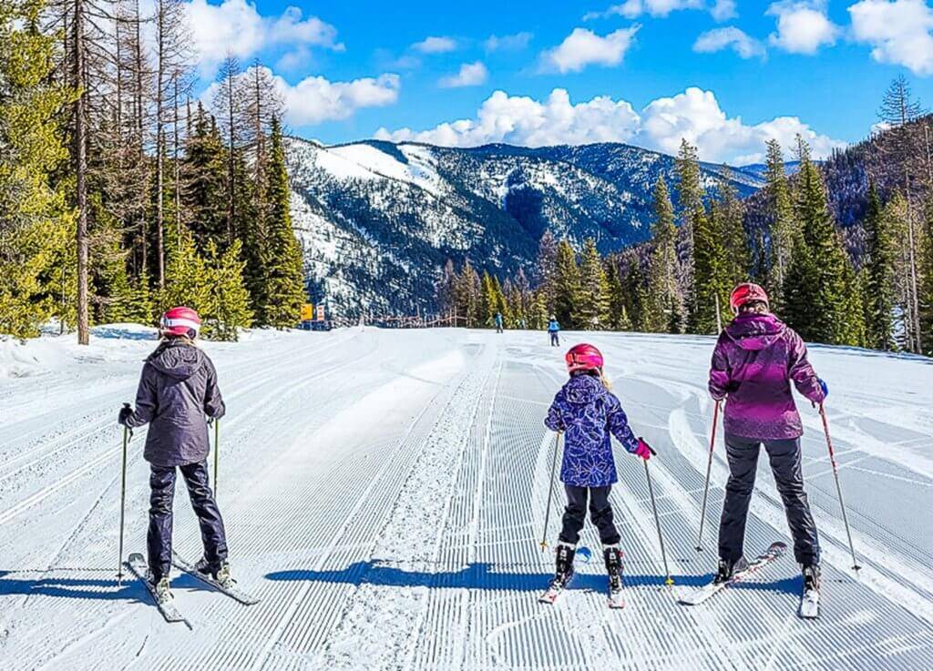 Two kids and an adult in snow gear ski a run at Lookout pass with a mountain in the distance.