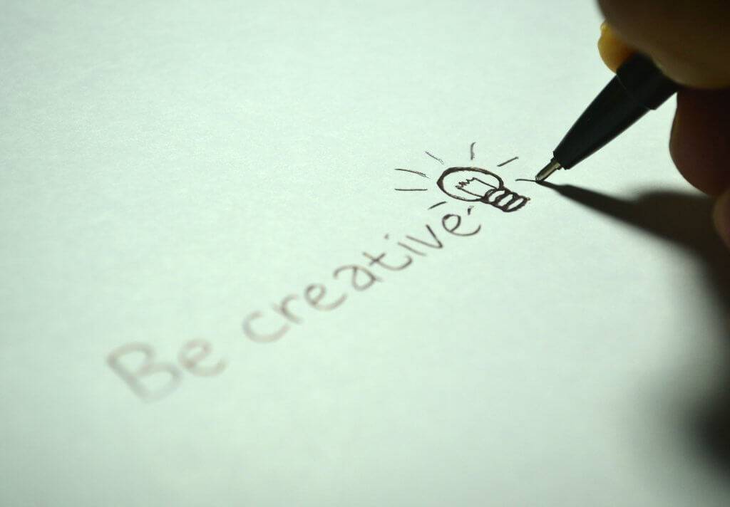 closeup of a hand holding a pen to paper and drawing a lightbulb beside the words be creative