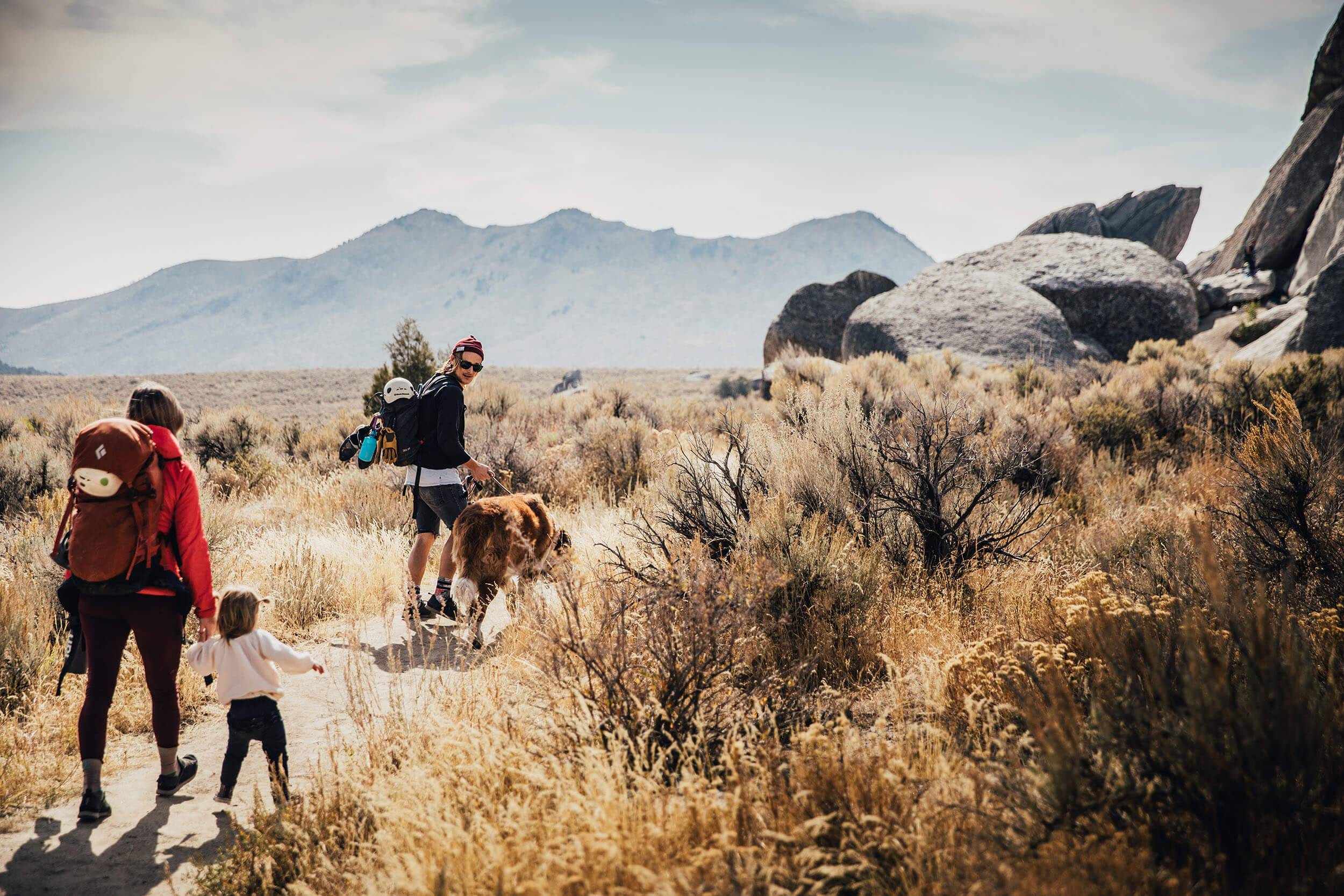 a family of three with their dog walking along a trail surrounded by desert shrubs with rock formations in the background