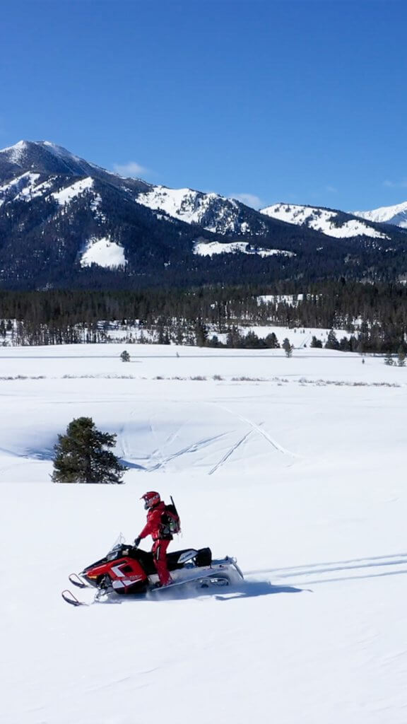 Two people riding across snowy backcountry at Smiley Creek on red snowmobiles with mountains behind them. 