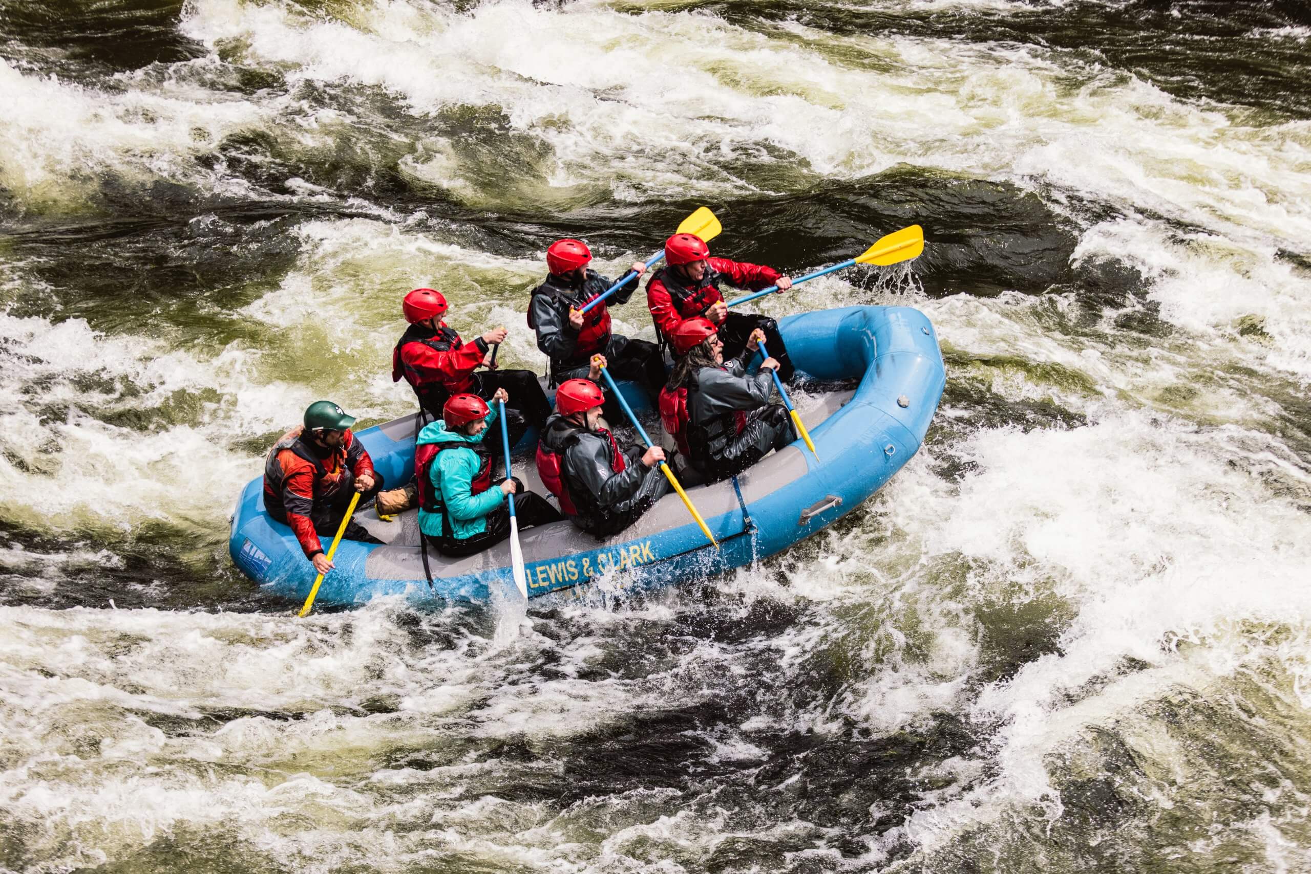 Seven people with red helmets sit in a blue raft for whitewater rafting.