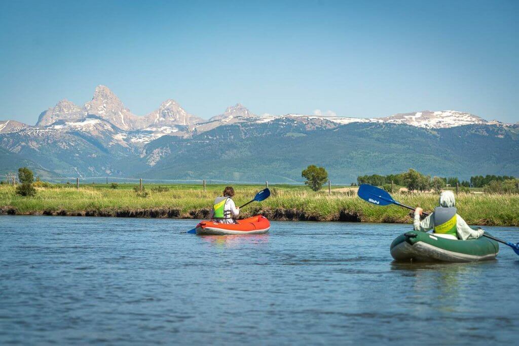Two people in inflatable kayaks paddling the Teton River with Teton Mountain Range in the background.
