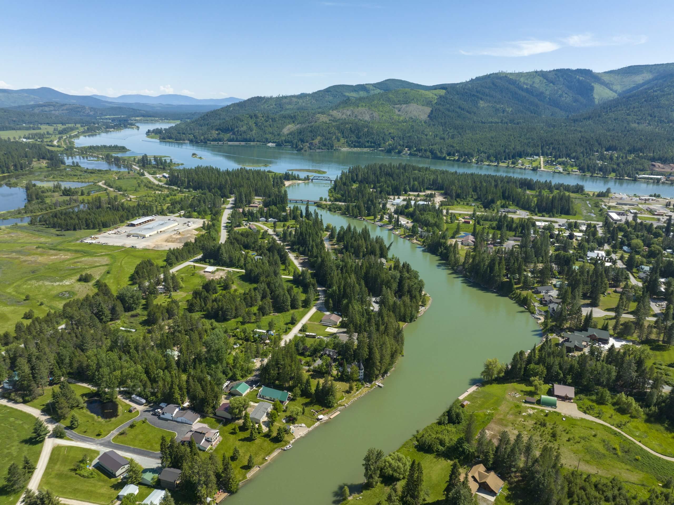 Aerial view of a river running through the town of Priest River, with tree covered mountains in the distance.