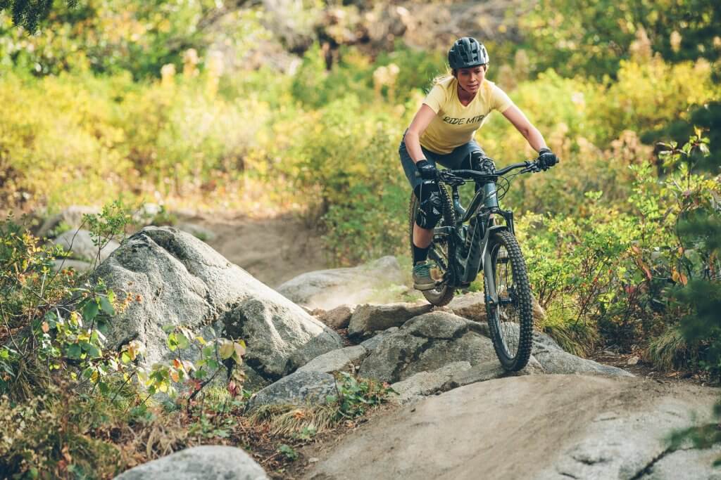 mountain biker, April Zastrow, wears a yellow shirt and rides over some rocks on her mountain bike