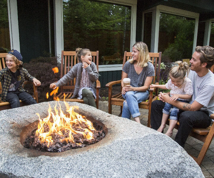 A group of two adults and three children sit around a lit fire pit laughing.