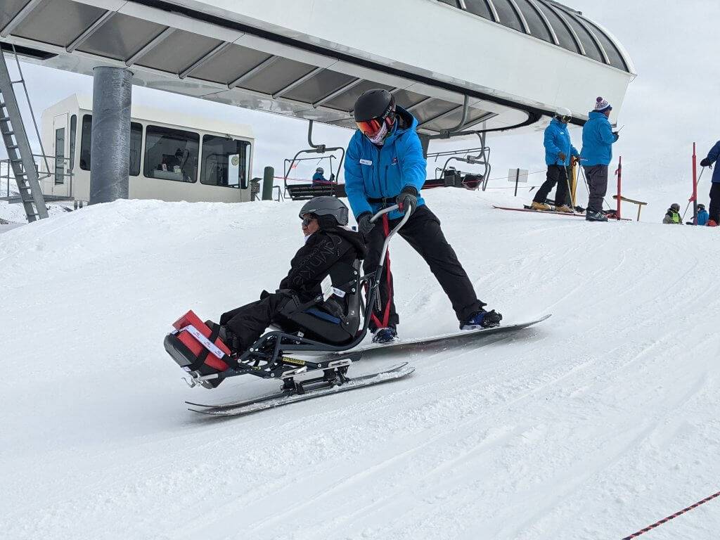 A ski instructor on a snowboard behind a skier in a sit ski helping them down the slope near the lift.