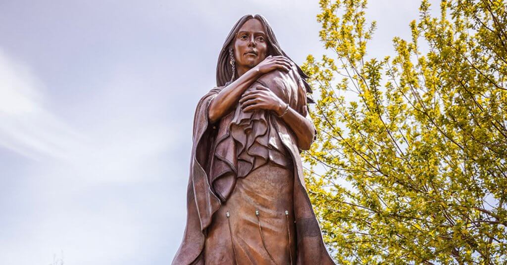 A bronze statue of Sacajawea holding a baby, with trees in the background.