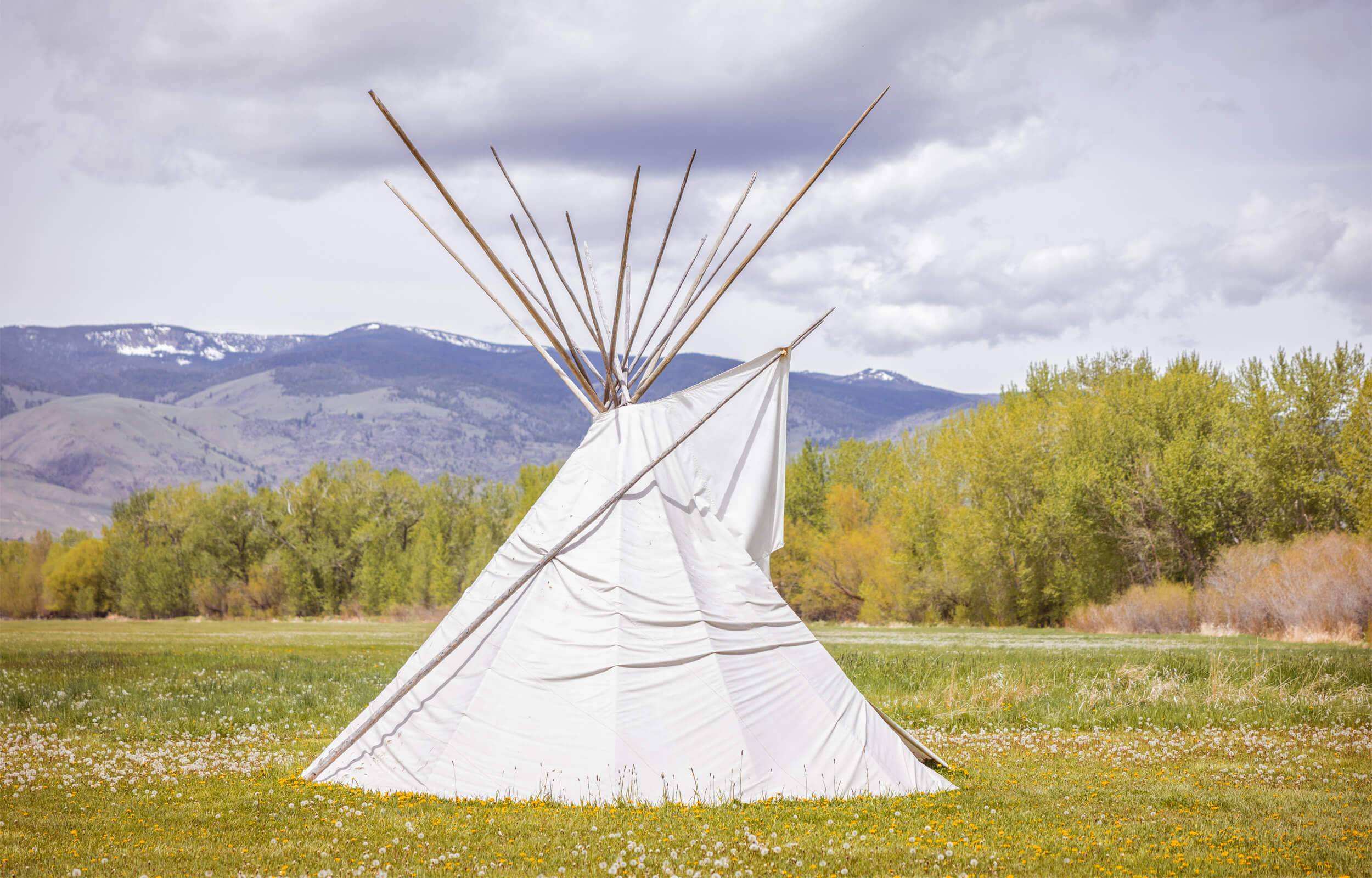 A white teepee in an open field, with trees and mountains in the distance, at the Sacajawea Interpretive, Cultural, and Educational Center.