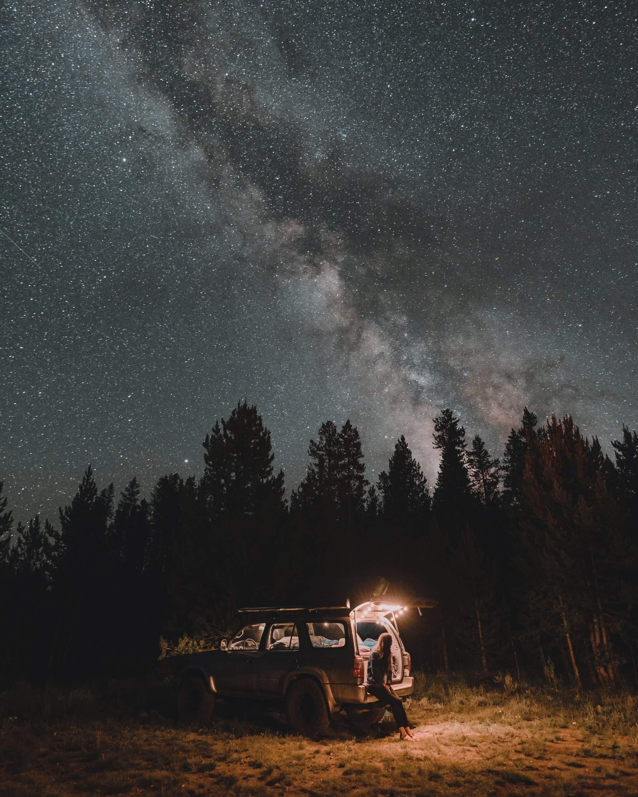 a person leaning against an suv amidst a forest under a starry sky