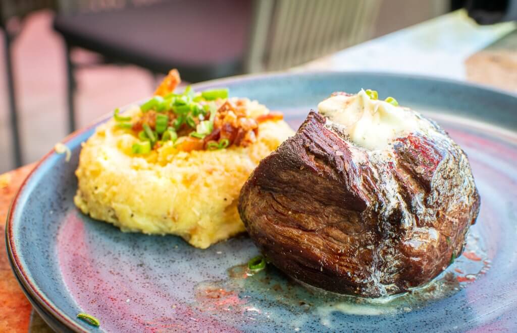 A steak sits at the center of a blueish plate with a serving of garnished mashed potatoes off to the side.
