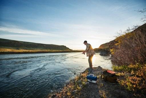 A man fishing along a river at sunset.