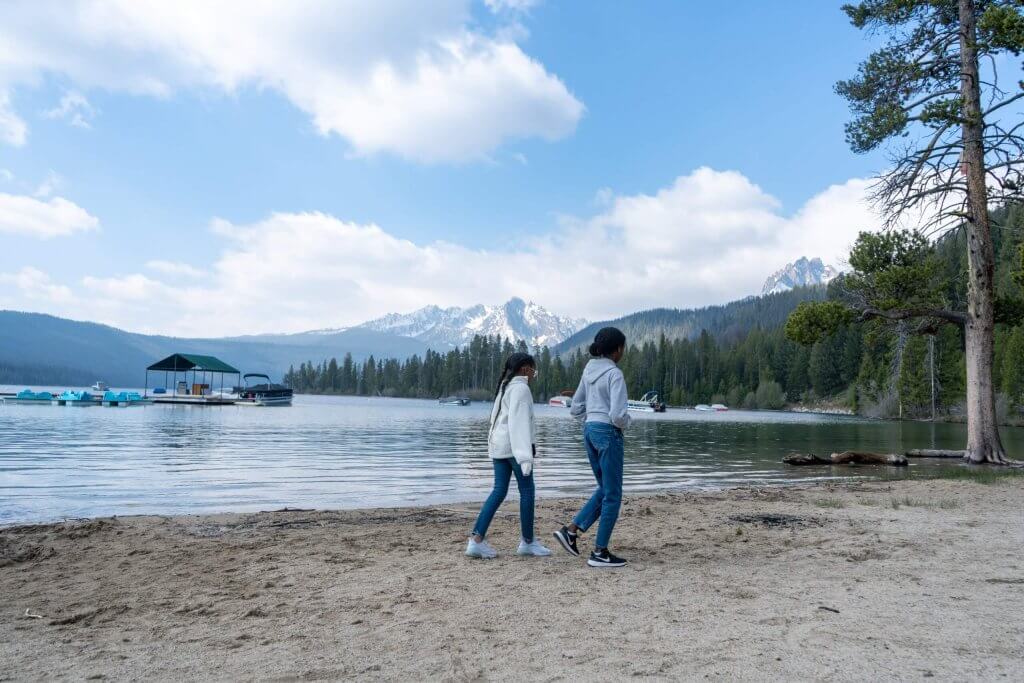 Two girls walk along the sandy shoreline of Redfish Lake with the Sawtooth Mountains and forest in the background.