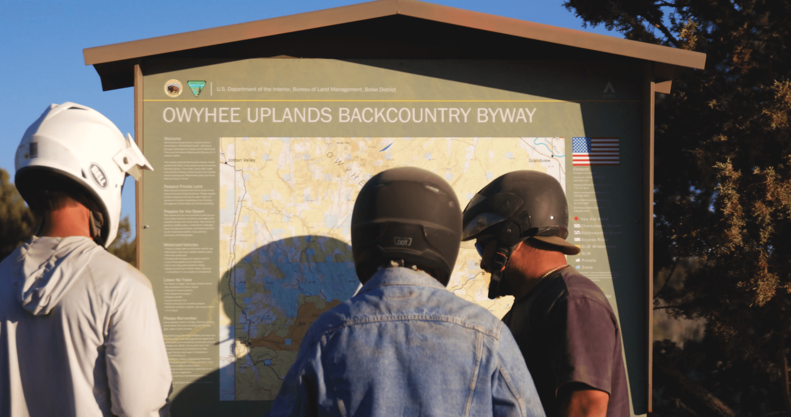 three people wearing helmets, looking at a Owyhee Uplands Backcountry Byway sign